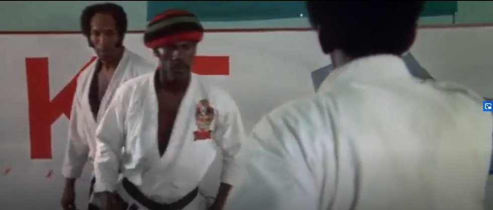 Three martial artists in white uniforms face each other in a dojo. One wears a colorful hat. Red text "KT" visible on the wall behind them.