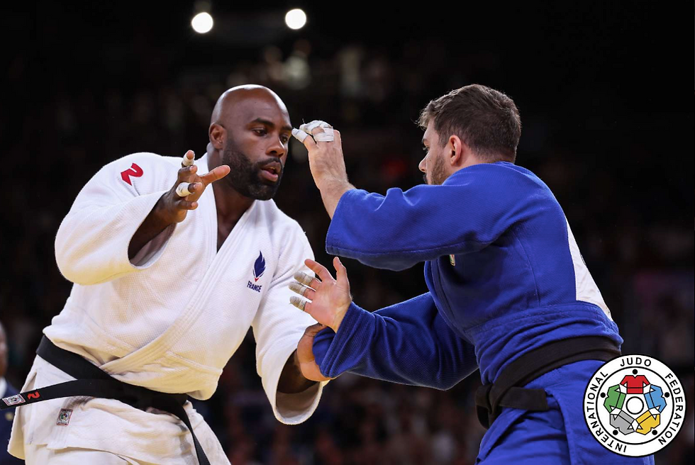 Two judokas in white and blue gis engage in a match. The setting is a sports arena with dim lighting. "International Judo Federation" logo visible.