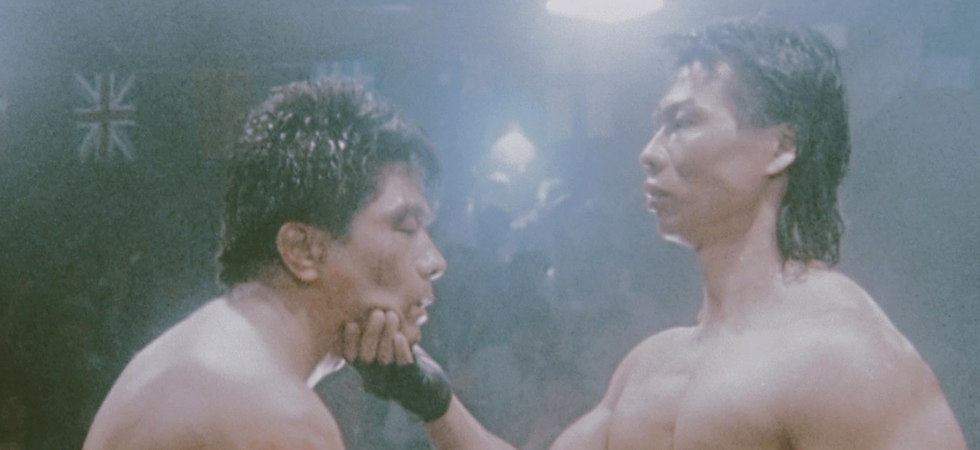 Two shirtless men face off in a dimly lit room, one raising the other's chin. A British flag is seen in the background. Tense atmosphere.
