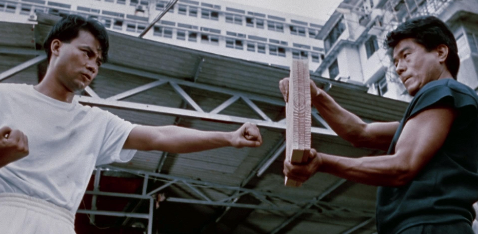 Two men outdoors, one in white punches a wooden board held by the other in black. Tense mood, industrial backdrop, and high-rise buildings.