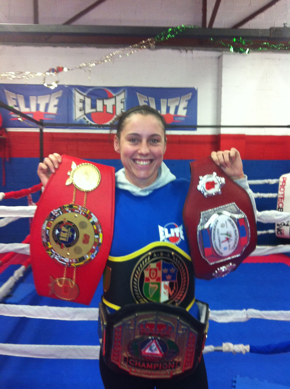 Smiling person holding two championship belts in a boxing ring. Blue and red gym with "ELITE" banners in the background.