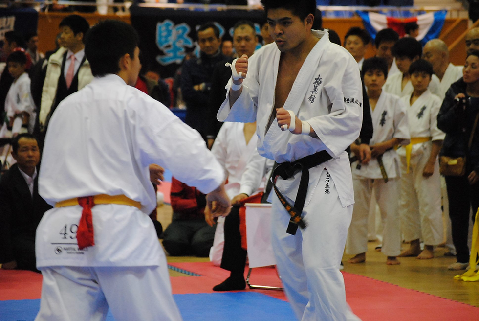 Two martial artists spar in white uniforms with belts in a crowded gym. Spectators watch intently. Mood is focused and intense.