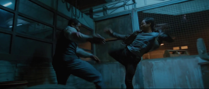 Two people engaged in a fight in a dimly lit, industrial setting. One aims a high kick at the other. The scene has a tense, action-packed mood.