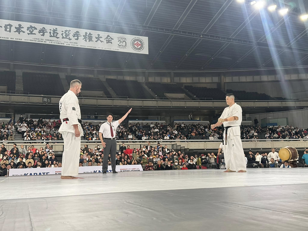 Two karate competitors face off in a packed arena. A referee raises his arm, signaling. Bright lights shine down on the white mat.