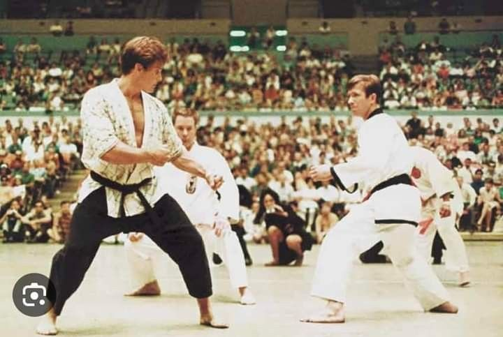 Two men in martial arts uniforms spar in an arena with a large crowd. Both wear white gis and black belts, focused and poised.