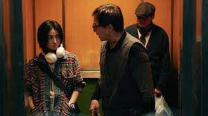 Two people converse in an elevator with wood paneling. The woman wears headphones and plaid, the man gestures while speaking. A third person stands quietly.