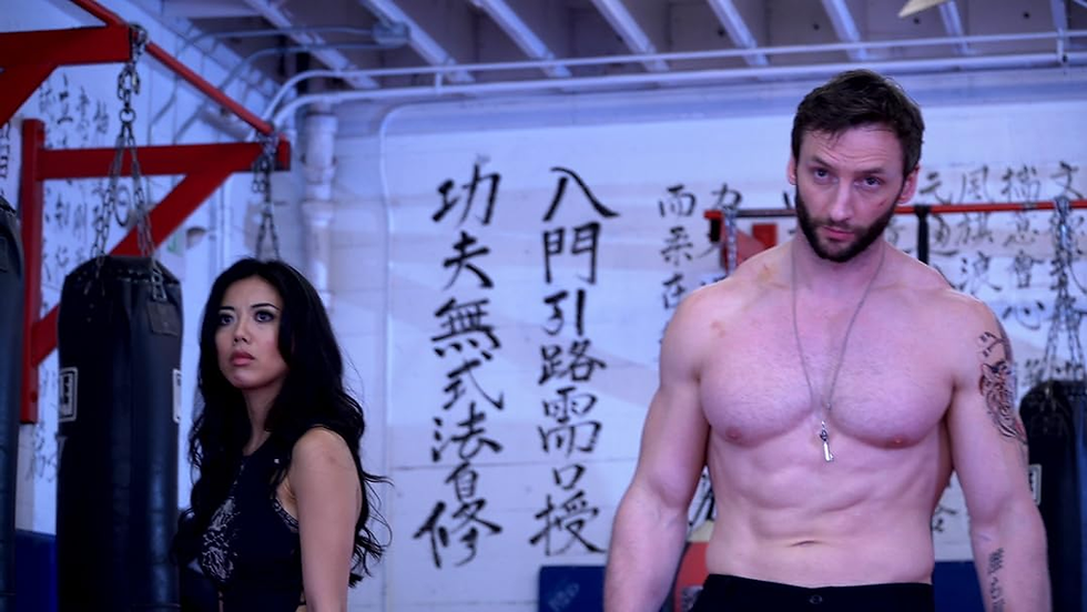 Man and woman in a gym with punching bags. The man is shirtless with tattoos, looking serious. Asian calligraphy decorates the wall.