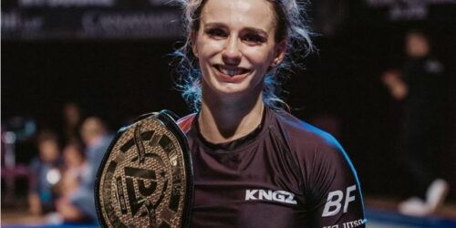Smiling person holds a championship belt, wearing a black KNGZ shirt. Background is blurred, creating a focused, triumphant atmosphere.