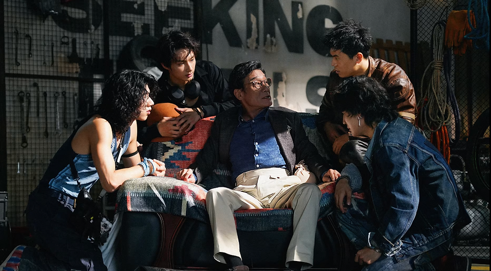 Five people in a dimly lit room, four standing and leaning in towards an older man sitting on a chair with a colorful blanket.